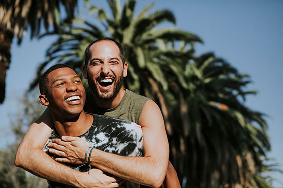 Same sex couple laugh outdoors with palm trees behind.